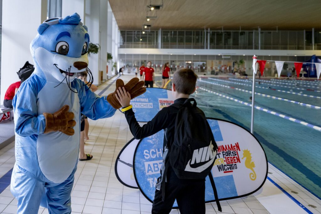 Das Maskottchen Otto des Deutschen Schwimm-Verbandes klatscht bei den Schwimmabzeichentagen 2026 mit einem Kind ab