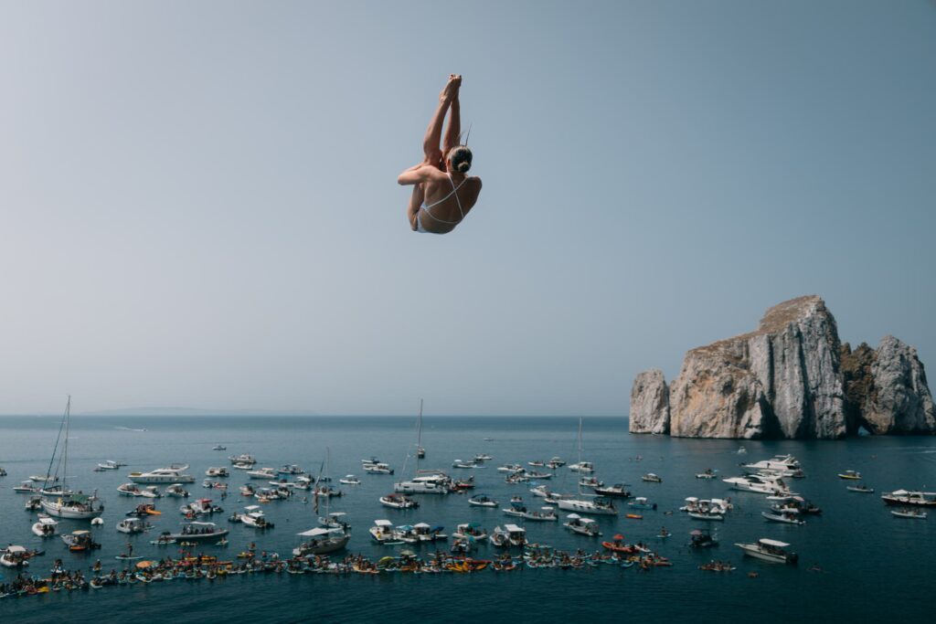 Eine Klippenspringerin beim Weltcup im High Diving 2026 in Porto Flavia auf Sardinien: Im Hintergrund sind Klippen und zahlreiche Boote auf dem Meer zu sehen, von denen aus die Menschen den Wettkampf verfolgen
