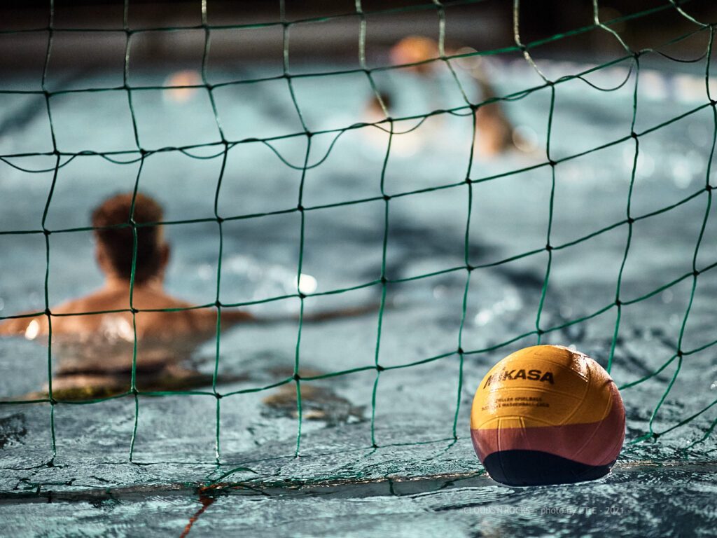 Symbolfoto vom Wasserballtraining: Ein Wasserball schwimmt im Tor, im Hintergrund ist der Torhüter zu sehen
