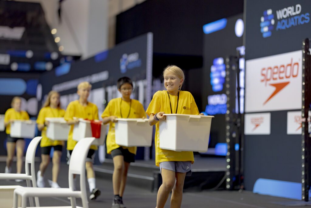 Kinder sind als Volunteers beim Schwimm-Weltcup 2023 in Berlin tätig und tragen die Körbe mit den Klamotten der Athleten
